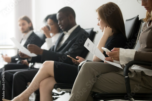 Multi-ethnic applicants sitting in queue preparing for interview, black and white vacancy candidates waiting on chairs holding resume using smartphones, human resources, hiring and job search concept