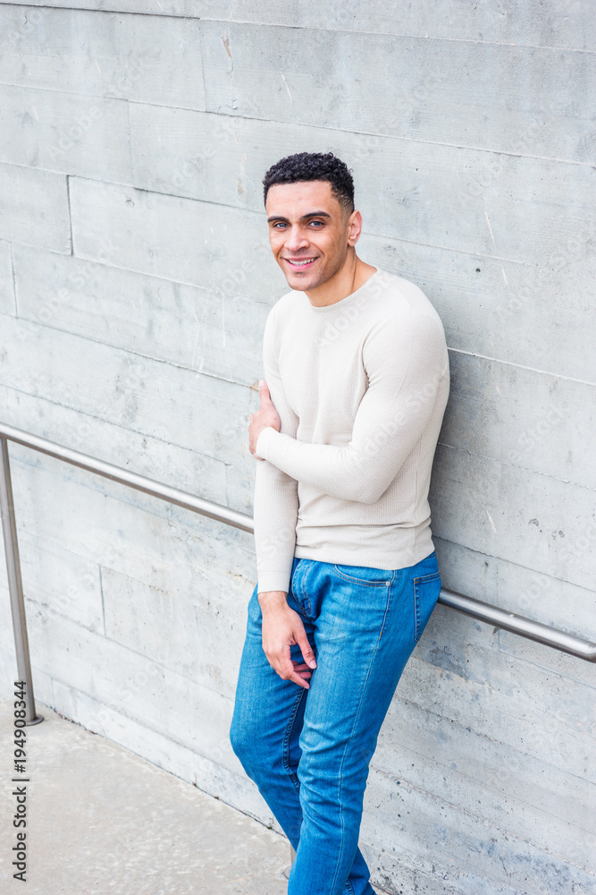 Young handsome American Man wearing light gray collarless, knit pullover sweater, blue jeans, standing by wall in New York in spring day, crossing arm, looking up, smiling.. Young handsome American Man wearing light gray collarless, knit pullover sweater, blue jeans, standing by wall in New York in spring day, crossing arm, looking up, smiling..