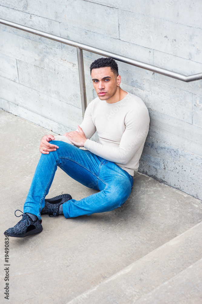 Young American Man wearing light gray knit sweater, blue jeans, black  sneakers, sitting on ground by wall in corner of street in New York,  looking up, thinking, lost in thought.. Stock Photo