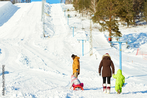 Rear view of parents with little kids going towards ski slope on beautiful winter day, father dragging sled with his daughter