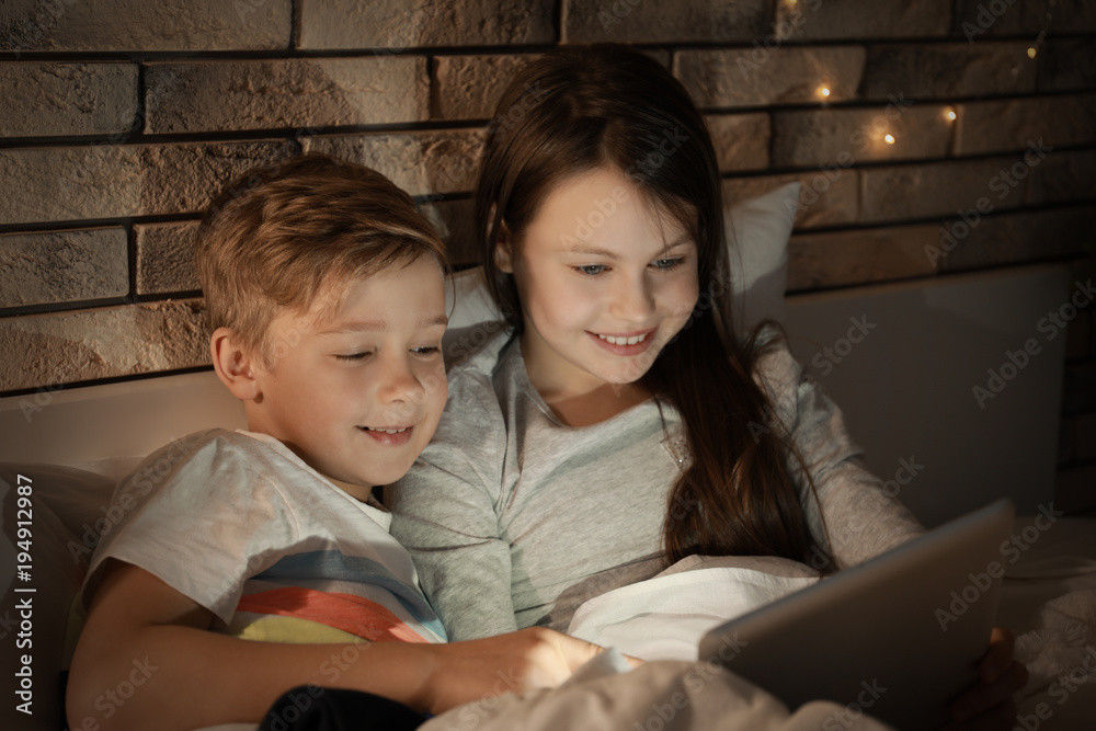 Cute little brother and sister reading bedtime story from tablet computer in evening Cute little brother and sister reading bedtime story from tablet computer in evening