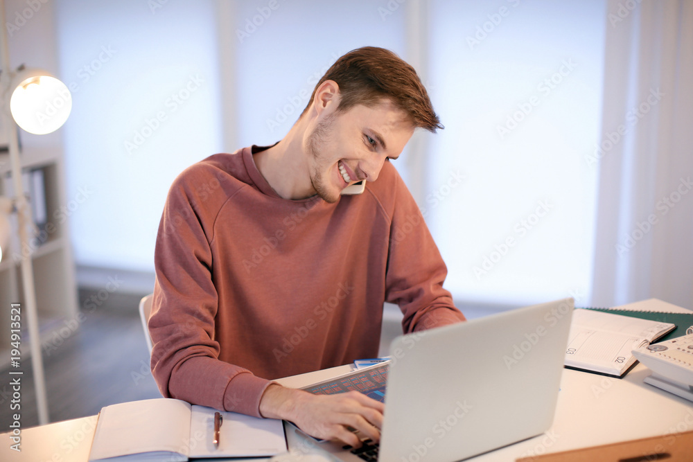 Young man talking on phone while working with laptop indoors Young man talking on phone while working with laptop indoors
