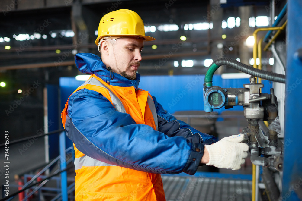 Profile view of confident bearded worker wearing hardhat and reflective vest standing at spacious production department of modern plant and adjusting equipment, portrait shot Profile view of confident bearded worker wearing hardhat and reflective vest standing at spacious production department of modern plant and adjusting equipment, portrait shot