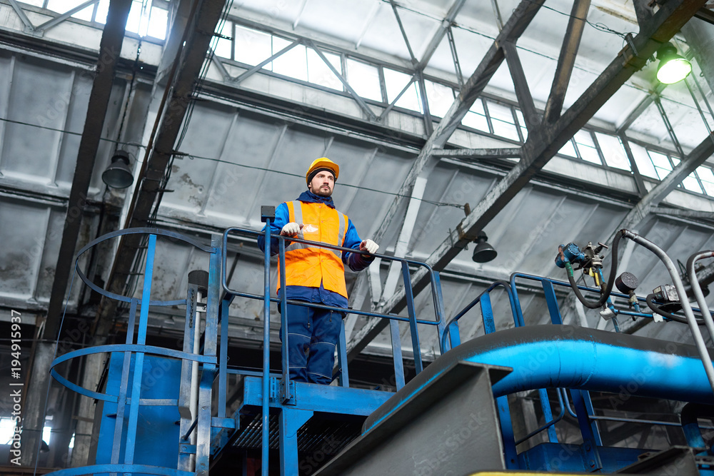Low angle view of handsome young worker wearing uniform and protective helmet looking at spacious production department of modern plant with concentration while controlling production process.