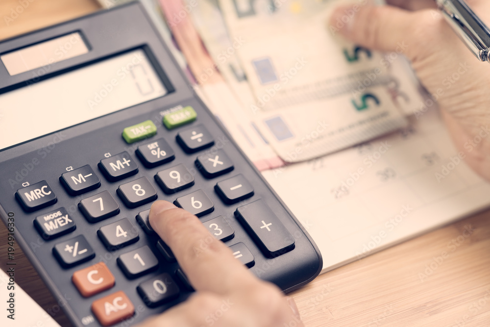 Cost calculation, payment day concept, hand put finger on calculator and other hand holding pen and pile of Euro banknotes with calendar on wood table, soft vintage effect Cost calculation, payment day concept, hand put finger on calculator and other hand holding pen and pile of Euro banknotes with calendar on wood table, soft vintage effect
