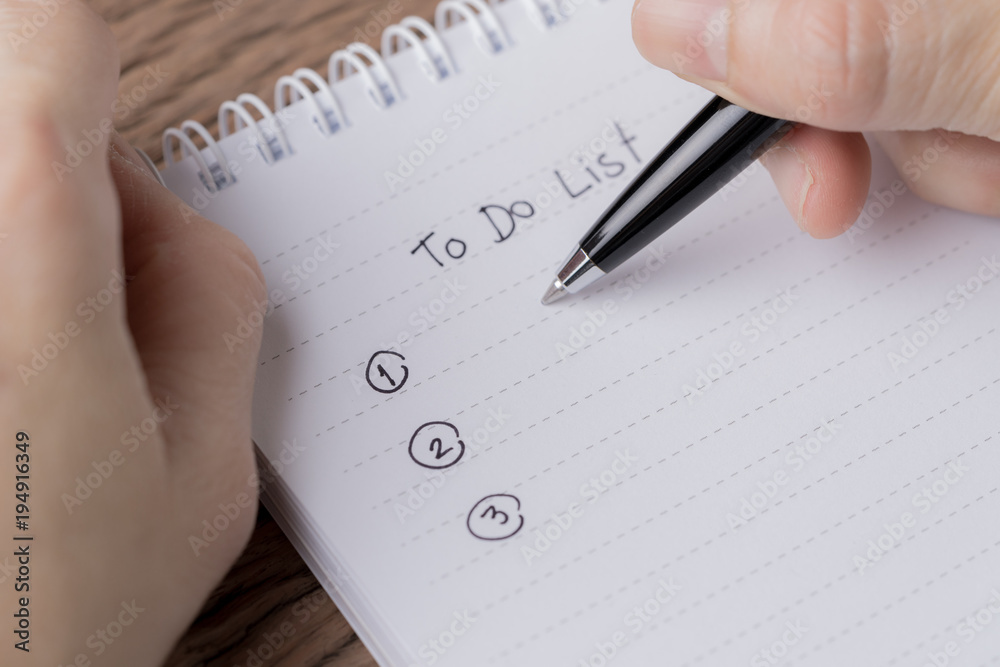 Female hand holding black pen writing To Do List prioritized by number on white paper notepad on wood table Female hand holding black pen writing To Do List prioritized by number on white paper notepad on wood table