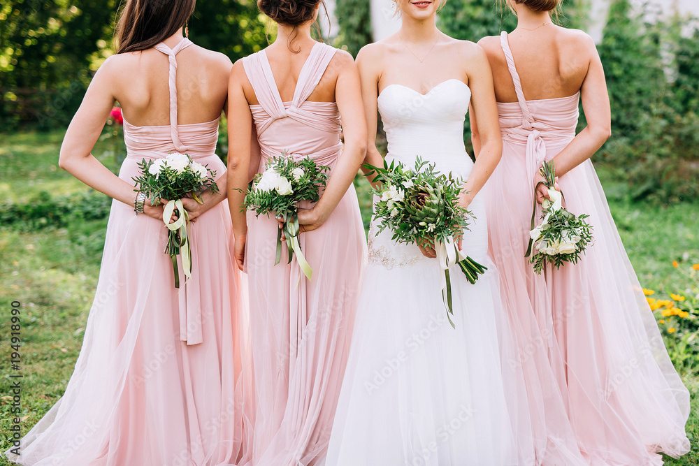 three bridesmaids in powdery dresses transformers with bouquets in hands stand with their backs near the bride in a white dress with a wedding bouquet in her hand on a green lawn - obrazy, fototapety, plakaty 
