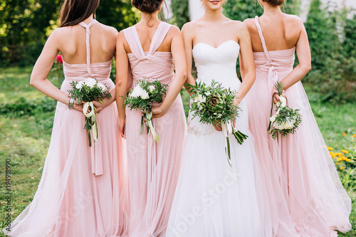 three bridesmaids in powdery dresses transformers with bouquets in hands stand with their backs near the bride in a white dress with a wedding bouquet in her hand on a green lawn photo