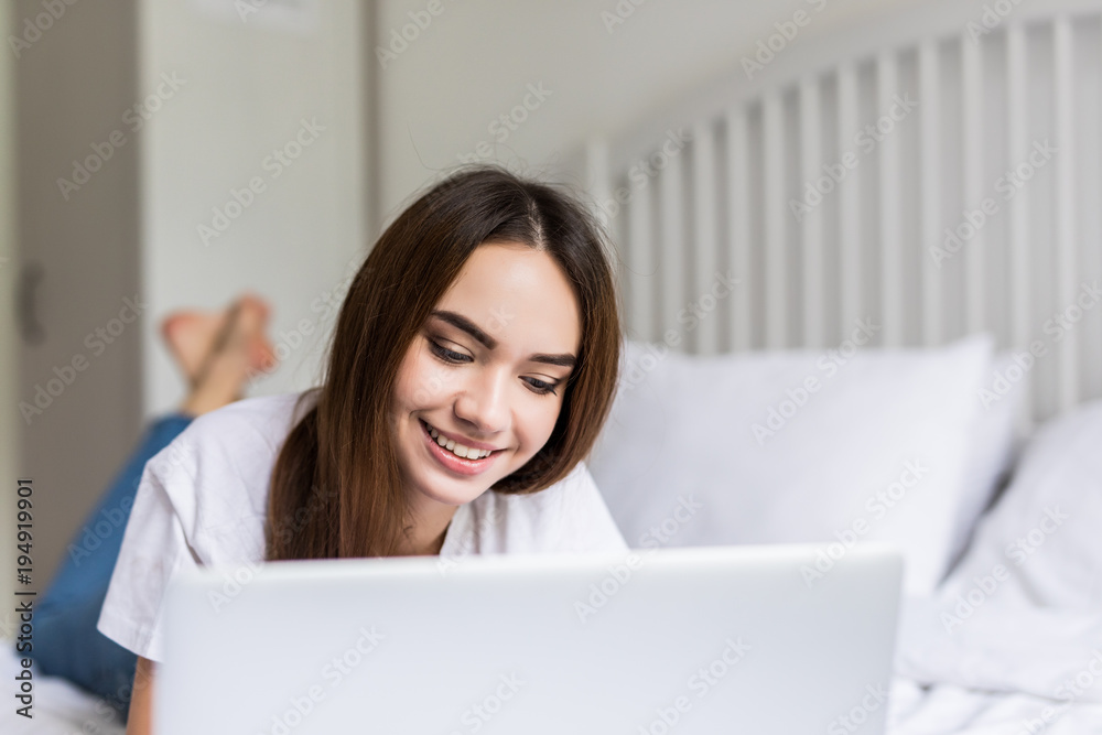 Smiling woman catching up on her social media as she relaxes in bed with a laptop computer on a lazy day Smiling woman catching up on her social media as she relaxes in bed with a laptop computer on a lazy day