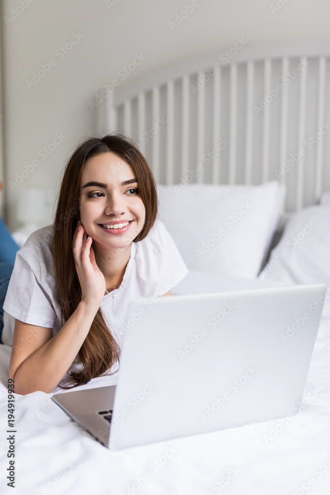 Smiling woman catching up on her social media as she relaxes in bed with a laptop computer on a lazy day Smiling woman catching up on her social media as she relaxes in bed with a laptop computer on a lazy day