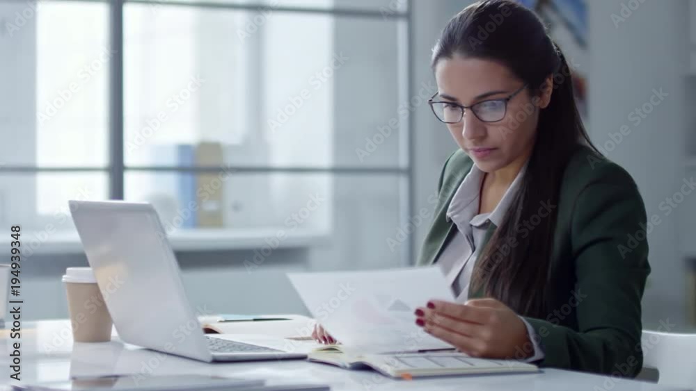 Tracking shot Latin businesswoman in eyeglasses typing on laptop at office desk and drinking coffee from disposal cup Tracking shot Latin businesswoman in eyeglasses typing on laptop at office desk and drinking coffee from disposal cup