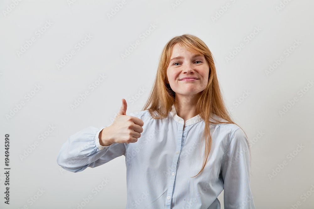 Studio portrait of pretty red-headed lady dressed in casual blue shirt smiling and showing big thumb up feeling enthusiastic and satisfied with a good quality. White background. Studio portrait of pretty red-headed lady dressed in casual blue shirt smiling and showing big thumb up feeling enthusiastic and satisfied with a good quality. White background.