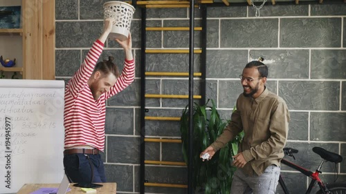 Two multiracial men have fun playing basketball game throwing paper in rubbish bin in modern office photo