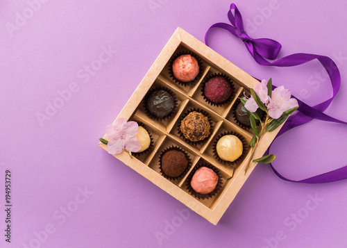 Assorted chocolates in a paper box with a satin purple ribbon on a bright background decorated with fresh flowers.