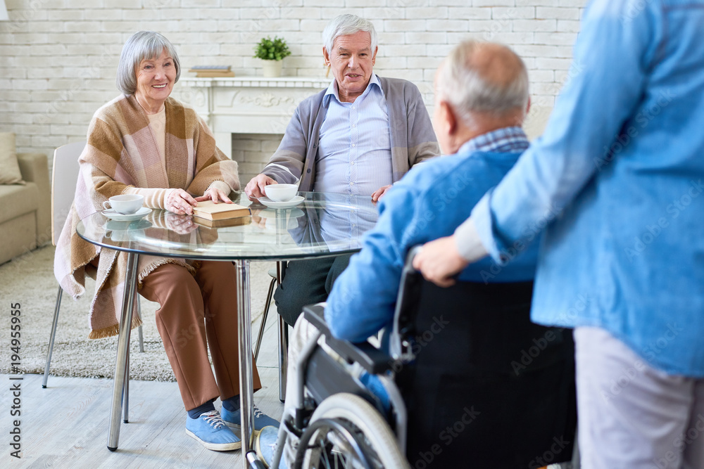 Obraz premium Portrait of nice senior couple smiling at friend in wheelchair coming to meet in retirement home living room