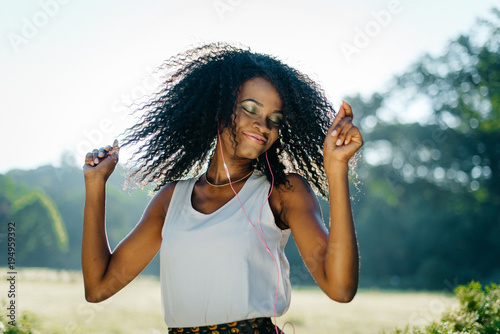 Emotional outdoor portrait. The charming young african girl with pretty smile and green eye shadows is shaking her curly hair while listening to music in her eyephones. photo