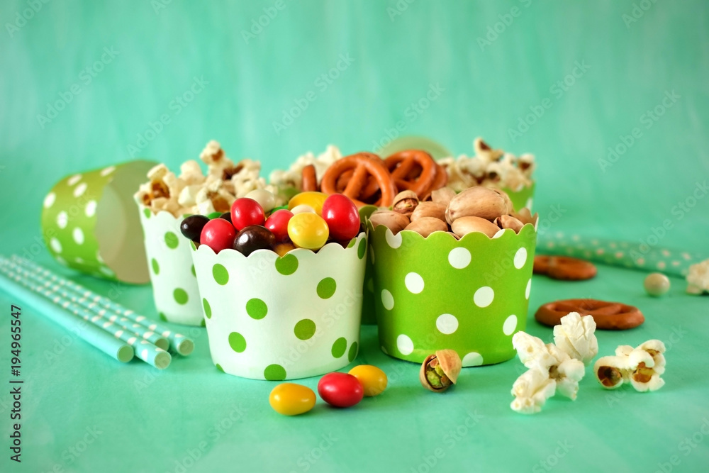 Popcorn, multicoloured drops, pretzels with salt and pistachio nuts in paper cups on green background. Snacks assortment