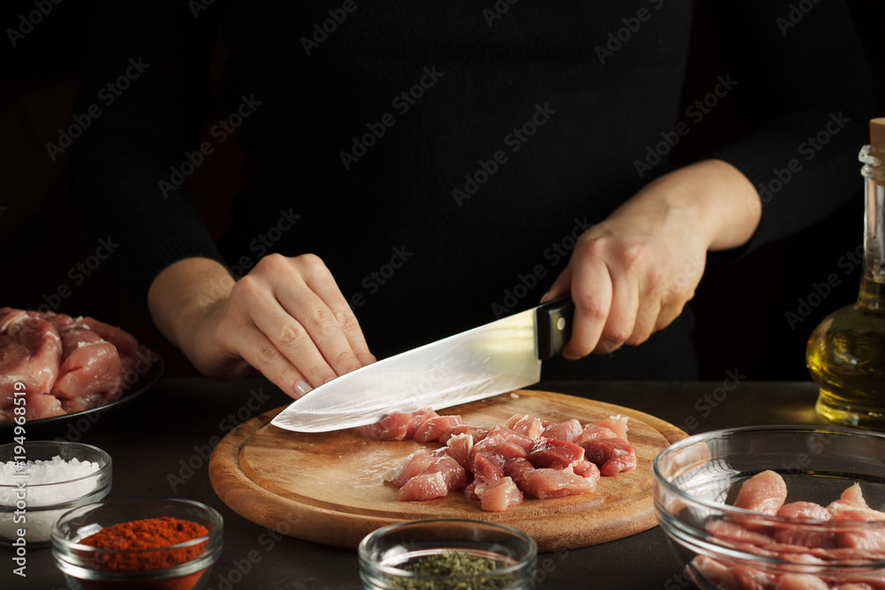 Female hands cut the raw meat into pieces on wooden board on dark table with seasoning and bottle of oil.