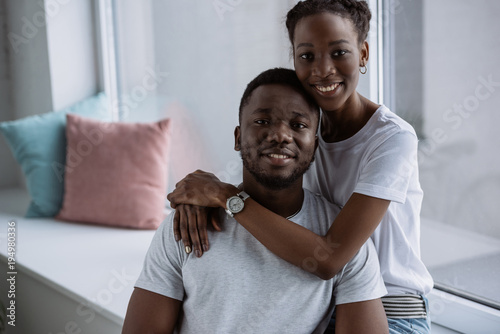happy young african american couple in white t-shirts smiling at camera at home