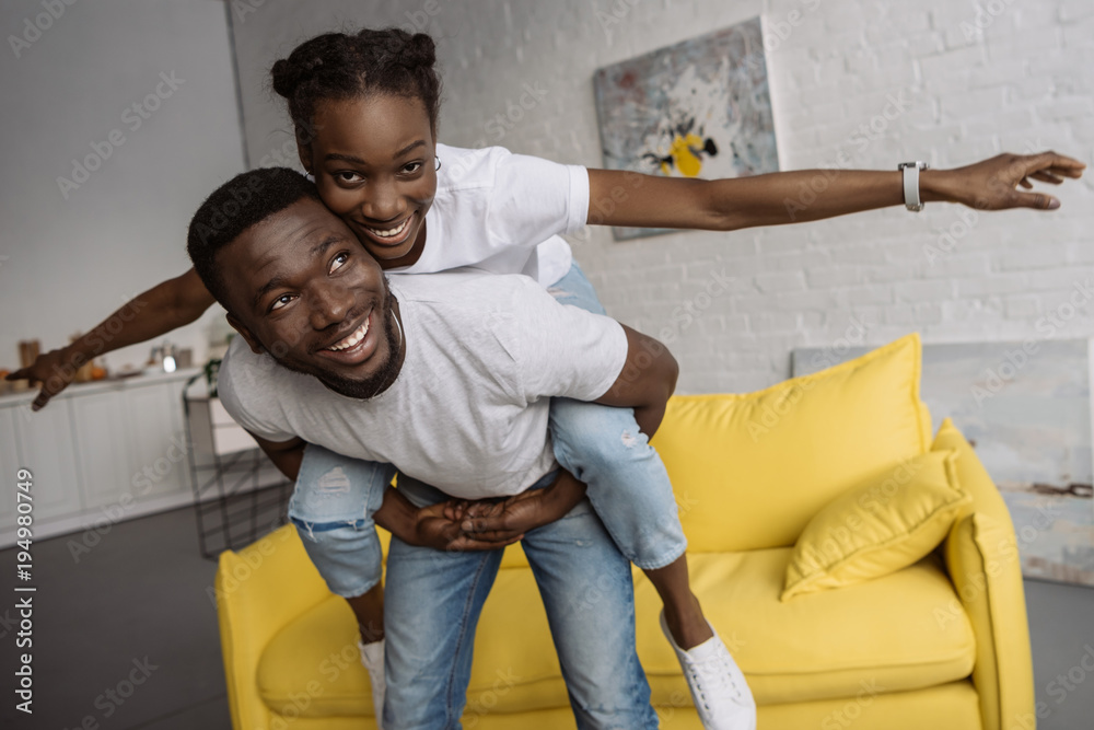 cheerful young african american couple piggybacking and having fun at home
