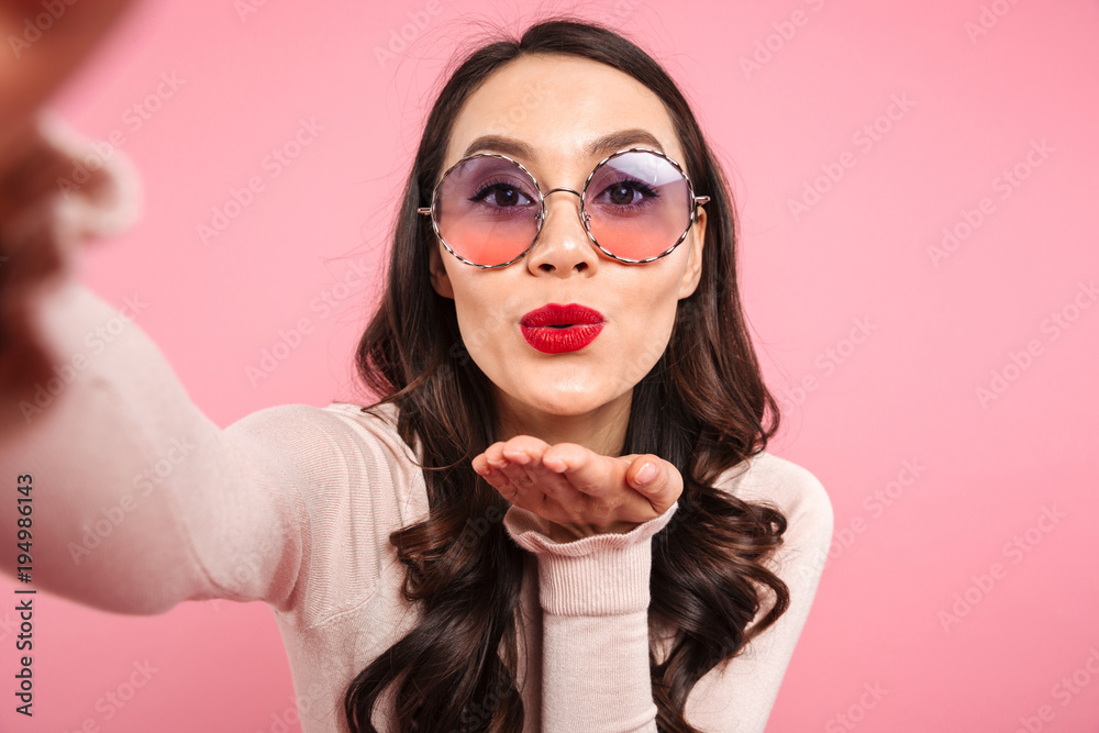 Cute lady in casual shirt with red lips wearing round sunglasses giving air kiss on camera while photographing herself, isolated over pink background Cute lady in casual shirt with red lips wearing round sunglasses giving air kiss on camera while photographing herself, isolated over pink background