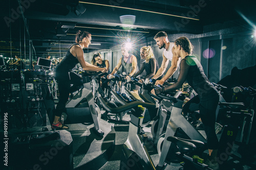 Sporty young women and men riding exercise bikes on cycling class with assistance of female instructor in the gym.