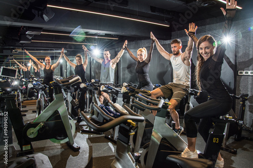 Fototapeta Naklejka Na Ścianę i Meble -  Group of fitness young women and men holding hands together and celebrating progress after successful cycling class in the gym.