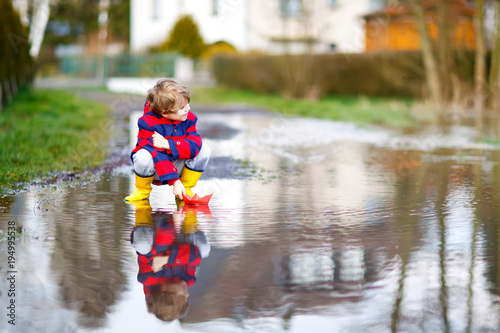 Happy little kid boy in yellow rain boots playing with paper ship boat by huge puddle on spring or autumn day