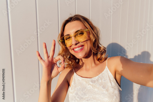 Cute funny girl in yellow eyeglasses smiling widely and showing a okay sign while taking a selfie  posing outdoors.