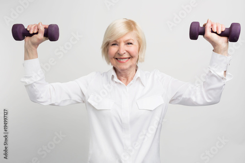 Cheerful madam is exercising with light weights. She does that because she has a careless retirement and much time to do that. Isolated on white background.
