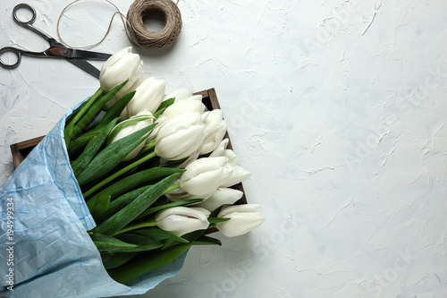 Fototapeta Naklejka Na Ścianę i Meble -  A bouquet of white tulips in blue wrapping paper in wooden box with scissors and twine on a white concrete background. Top view. Flat lay. Postcard for Easter, Mother's Day and Spring Holidays.