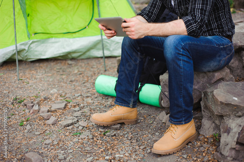 Traveler near a tent with a tablet in the evening. Rocky mountains background.