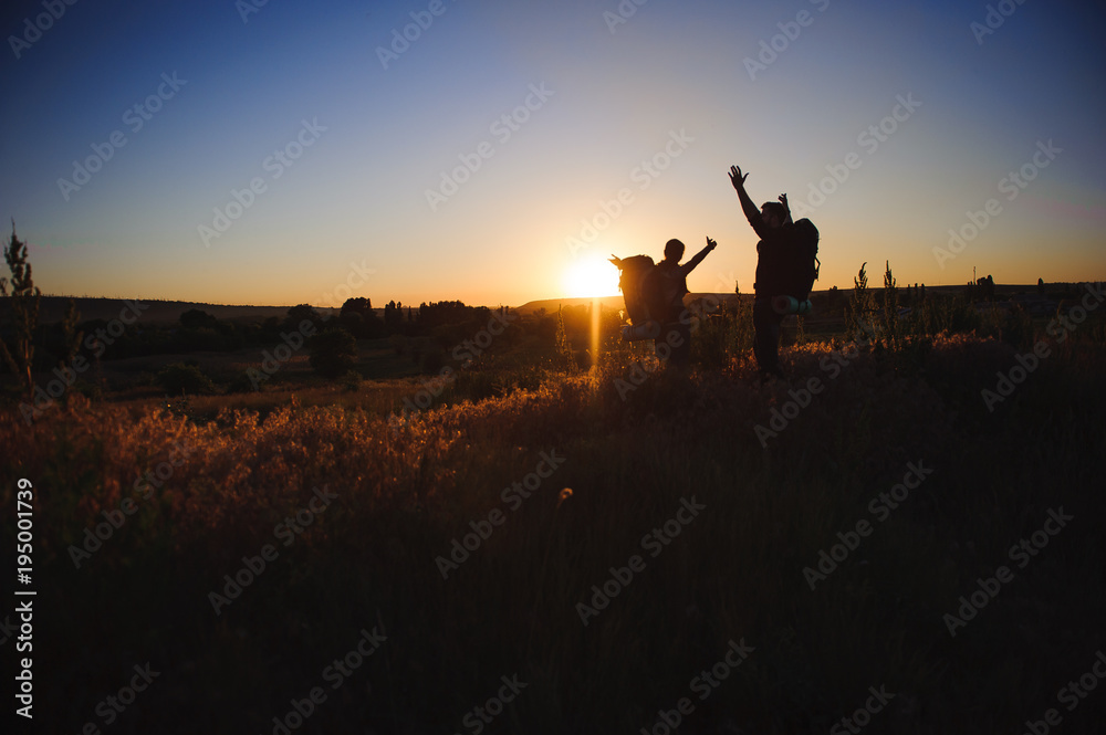 Fototapeta premium Silhouettes of two hikers with backpacks walking at sunset. Trekking and enjoying the sunset view
