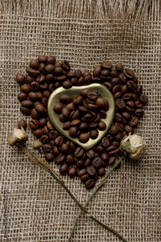 Flat lay of roasted coffee beans on a tablecloth with a golden heart shaped saucer and coffee mug. Cup of morning espresso and pressed roses. Romantic ideas