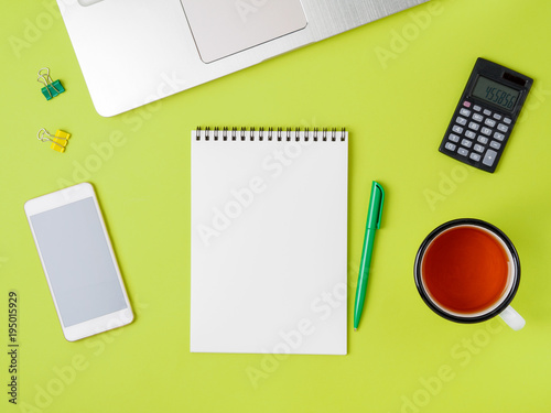 Modern creative bright green office desk with laptop, smartphone and other accessories. Blank Notepad page for text in the middle, top view.