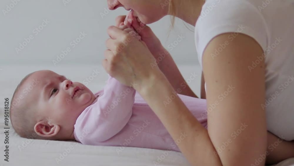 Mother kisses baby daughter feet while playing on a white bed Mother kisses baby daughter feet while playing on a white bed