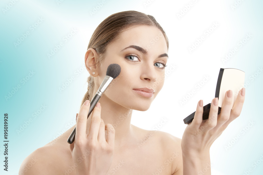 Fototapeta premium Close-up of a young woman holding brush in her hand and applying makeup. Isolated on light blue background.