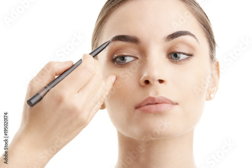 Studio shot of a beautiful young woman applying makeup to her eyebrow while standing at isolated background.