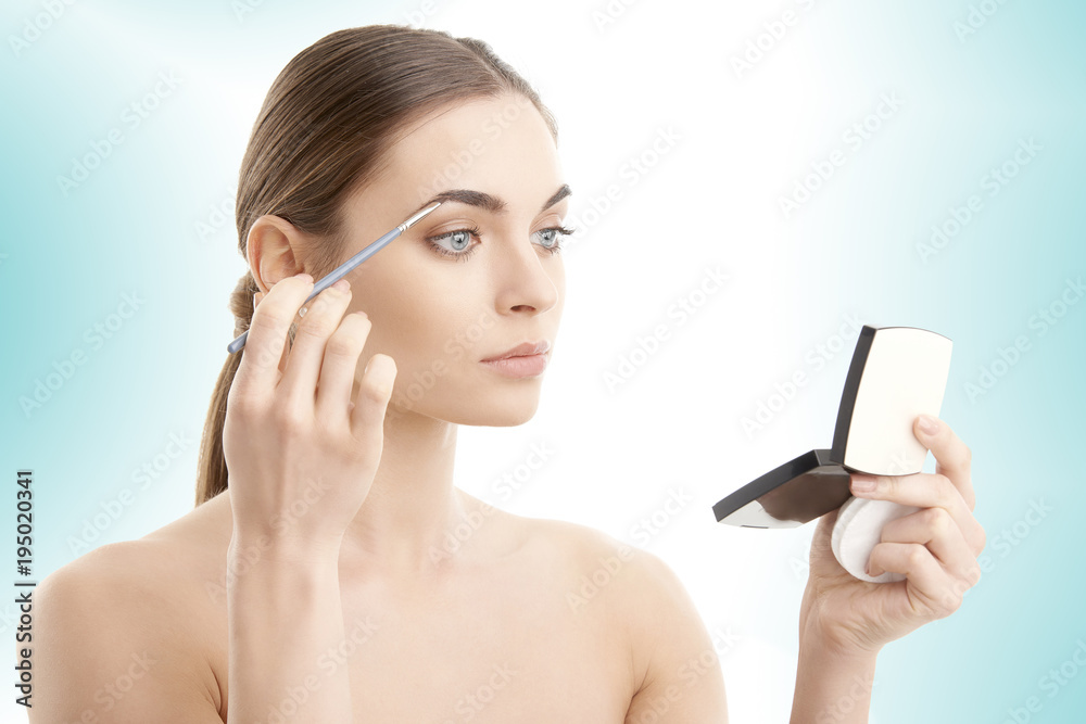 Studio shot of a beautiful young woman applying makeup to her eyebrow while standing at isolated background. Studio shot of a beautiful young woman applying makeup to her eyebrow while standing at isolated background.