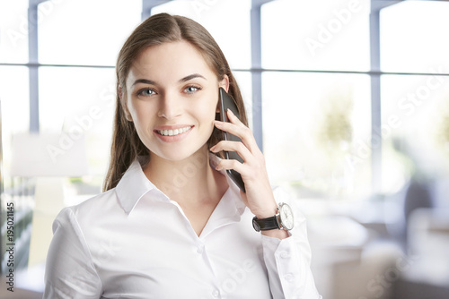 Young professional woman with her cellphone. Attractive young financial assistant businesswoman talking with somebody with her mobile phone while standing at the office. 