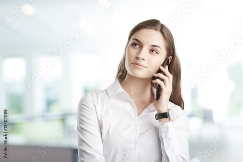 Young professional woman with her cellphone. Attractive young financial assistant businesswoman talking with somebody with her mobile phone while standing at the office. 
