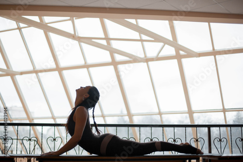 brunette woman practicing yoga concept, stretching in Cobra exercise, Bhujangasana pose on table top in front of large windows. working out, full length, wearing sportswear. photo