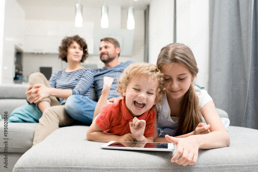 Portrait of brother and sister using digital tablet and laughing while lying on sofa in living room with parents watching TV in background, copy space Portrait of brother and sister using digital tablet and laughing while lying on sofa in living room with parents watching TV in background, copy space