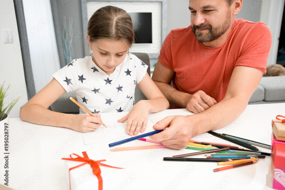 Portrait of bearded father helping little girl drawing handmade card for mothers day at home in modern living room Portrait of bearded father helping little girl drawing handmade card for mothers day at home in modern living room