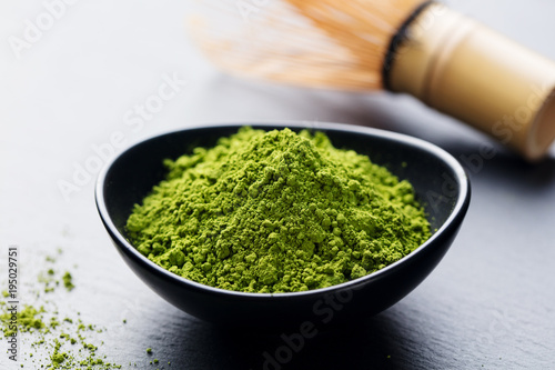 Matcha, green tea powder in black bowl with bamboo whisk on slate background. Copy space.
