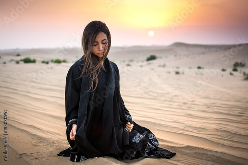 Beautiful young woman in black dress sitting on sand in the desert.