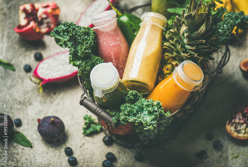 Colorful smoothies in bottles with fresh tropical fruit and greens in basket on grey concrete background, selective focus. Healthy, vegetarian, detox, dieting, clean eating breakfast food concept