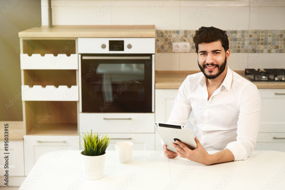 Handsome young boy with back wavy hair and neat beard using tablet in modern, light kitchen, wearing pristine white shirt