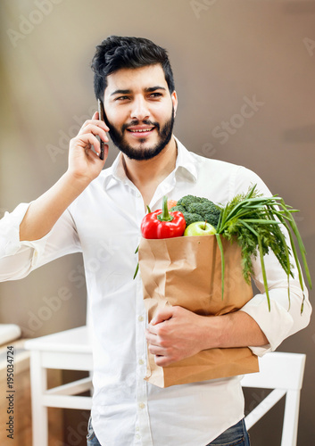 Handsome bearded man holding shopping bag with healthy, nutritious food talking by phone photo