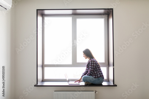 Beautiful young woman sitting on windowsill and using laptop against window